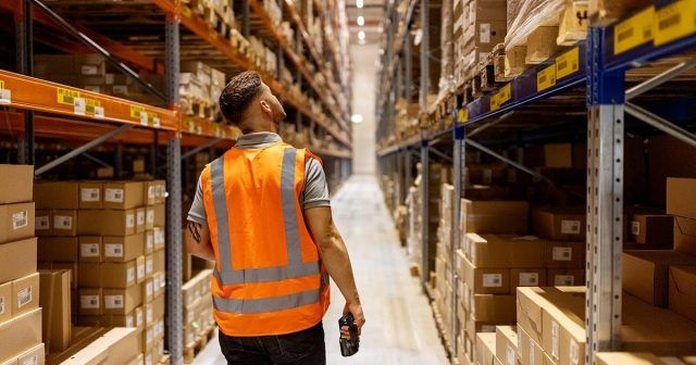 Man in reflective vest walking through a warehouse aisle, holding a device and looking up.