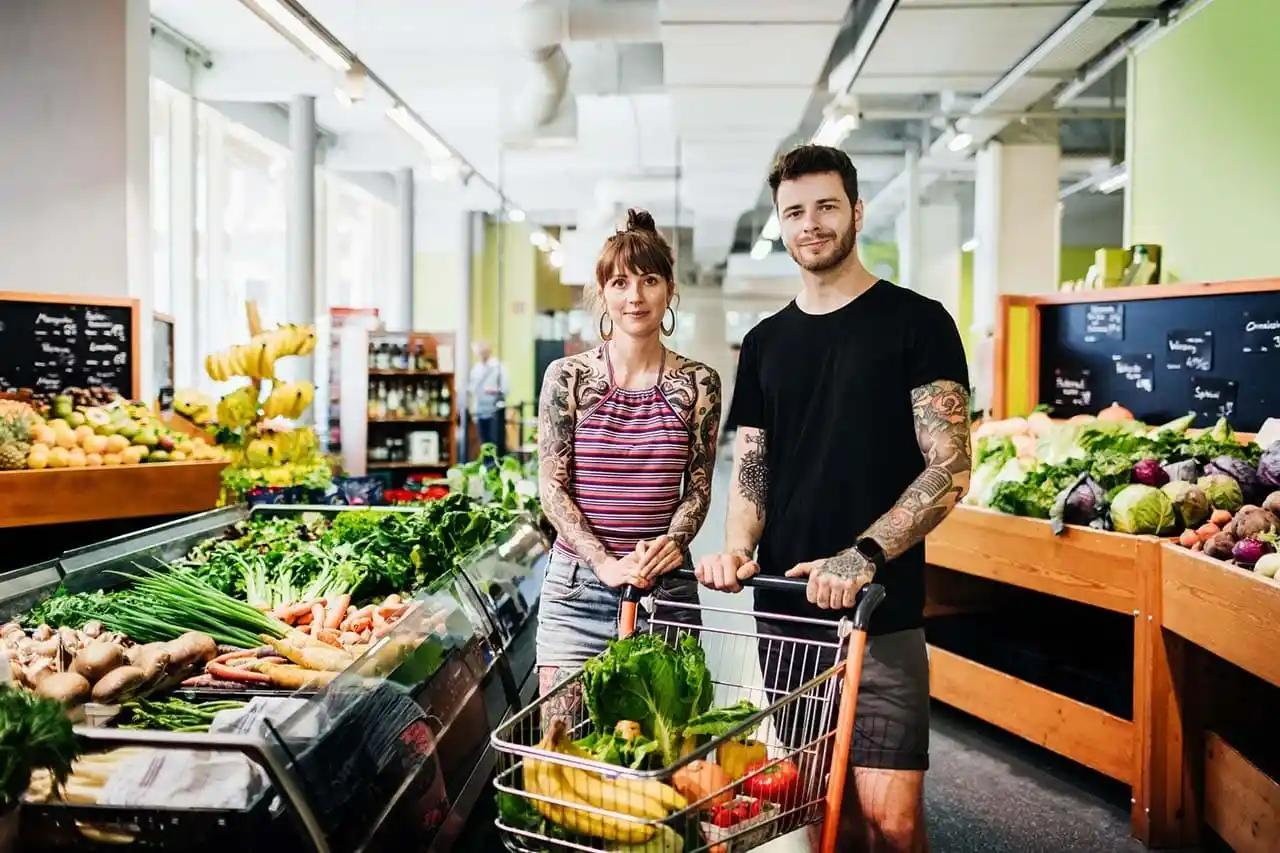 A man and woman stand in a grocery store beside a cart filled with fresh produce.