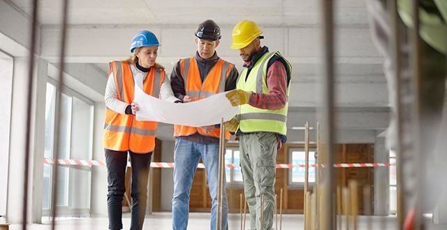 Three workers in safety vests and helmets examining a blueprint in an unfinished building.