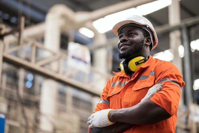 A construction worker in an orange safety uniform and white helmet stands confidently with arms crossed. He wears yellow headphones around his neck and gloves on his hands, embodying the meticulous standards of engineering insurance. Industrial equipment is visible in the blurred background.