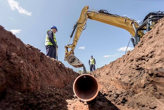 Construction workers beside an excavator digging a trench with a large pipe laid inside.