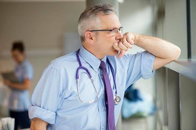 A male doctor in a blue shirt, and tie, with a stethoscope leans against a window, looking thoughtfully outside.