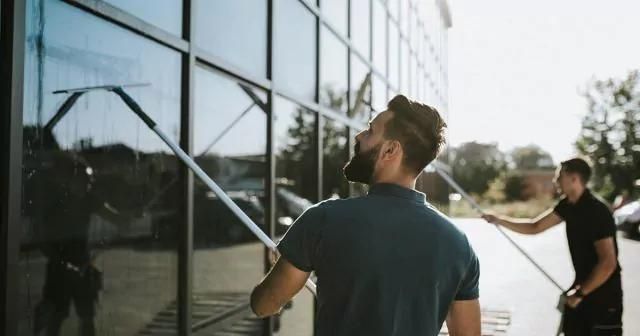 Two window cleaners using long squeegees on large glass windows, with trees and a clear sky in the background.