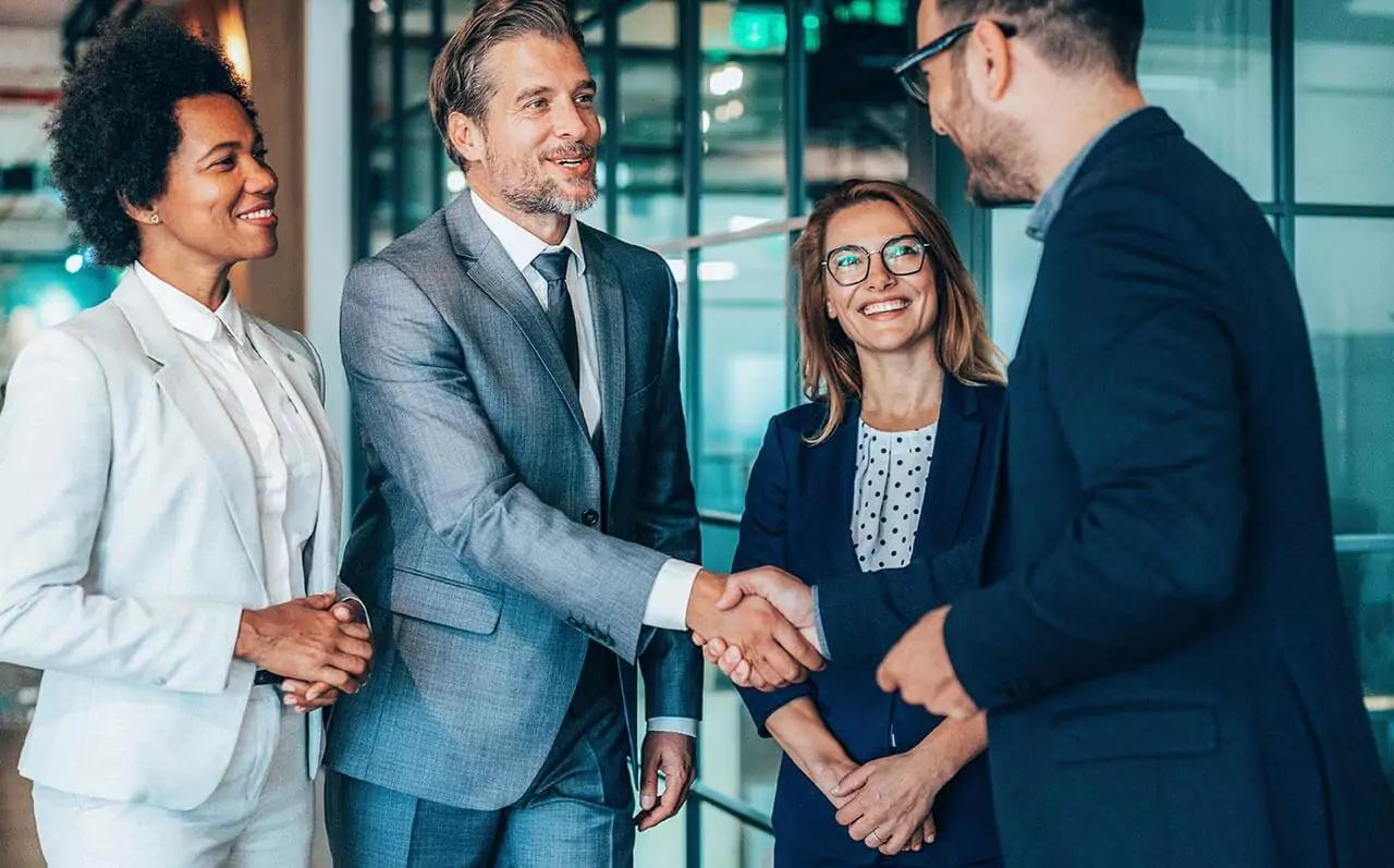 Four people in business attire shaking hands in hallway of modern office.