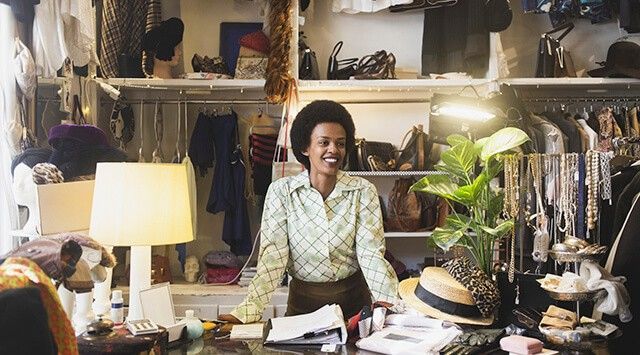 A retail charity store owner smiles behind a cluttered counter with vintage clothes and accessories.