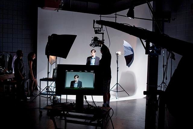 A person stands in front of a white backdrop, posing for a photoshoot. Studio lights, umbrellas, and other equipment surround them. A monitor displays the persons image, and silhouettes of crew members are visible.