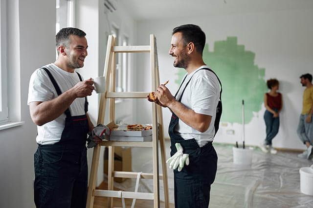 Two men in work overalls stand beside a wooden ladder, holding slices of pizza and a mug, and smiling at each other. In the background, two people near a partially painted wall appear to be talking. Paint supplies are scattered around the room.