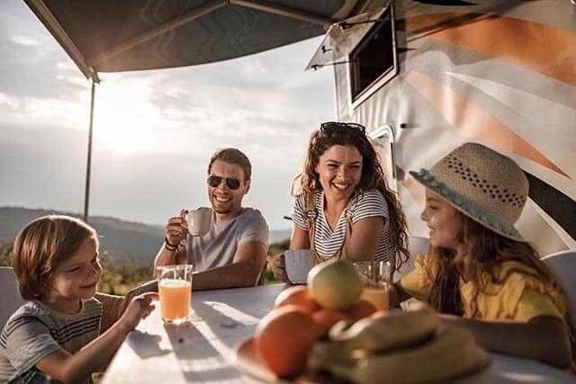 A family of four sits at an outdoor table beside a camper. They are smiling and enjoying drinks. There are fruits like bananas and apples on the table. The setting is sunny, with hills in the background, and an awning provides shade.
