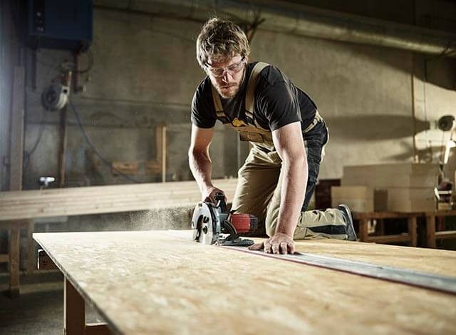 Person cutting a wooden board with a circular saw in a workshop, dust visible in the air.