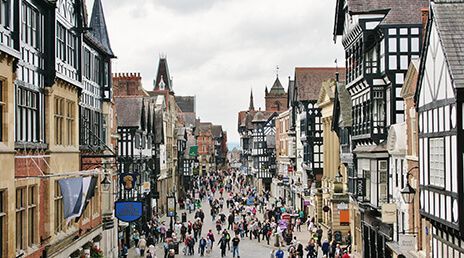 A bustling street scene in a historic town, featuring Tudor-style buildings with black and white timber framing. The street is filled with people walking, and the sky is overcast, adding a subdued atmosphere to the scene.