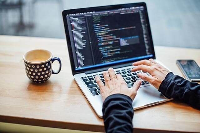 Person writing code on an open laptop in a modern workspace, with a mobile phone and mug filled with coffee nearby.