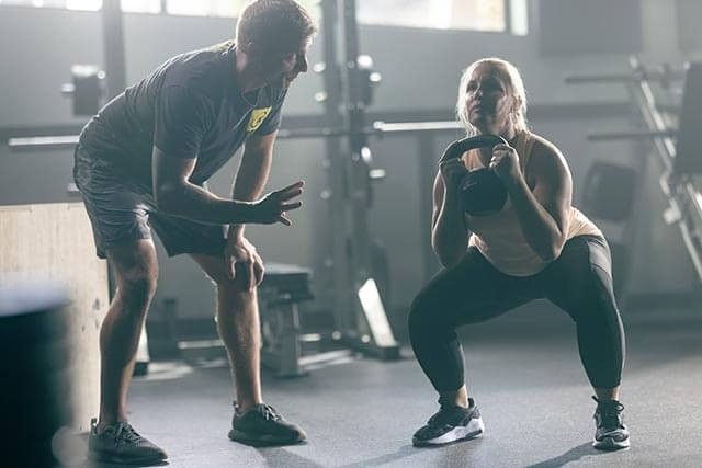 A woman executes a squat with a kettlebell in a well-equipped gym. Beside her, a man crouches, providing guidance. With weights and machines around, the gyms atmosphere highlights the need for proper training and gym insurance to ensure safety for all.