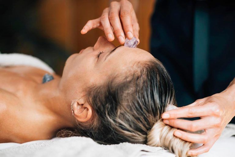 Person lying on a massage table, eyes closed, while another holds an amethyst crystal on their forehead.