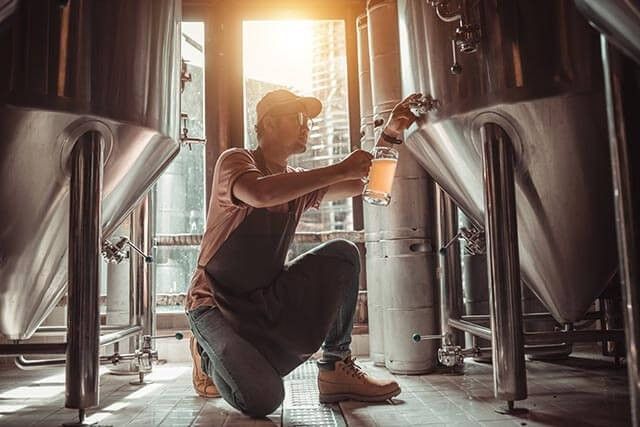 A person in a cap and apron kneels on the tiled floor of the brewery, carefully filling a glass from a tap on the large metal brewing tank. Sunlight streams through a window, casting warm light over this serene moment.
