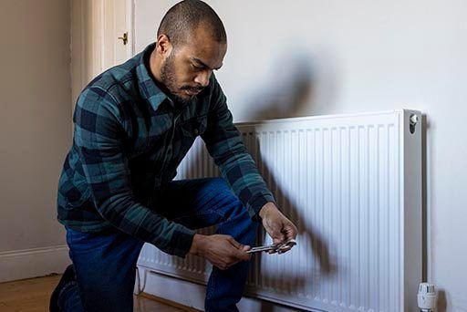 A person in a plaid shirt kneels while using a wrench to adjust a radiator in a room, perhaps mindful of block of flats insurance requirements. The plain wall behind contrasts the wooden floor, and an electrical outlet is visible near the white radiator.