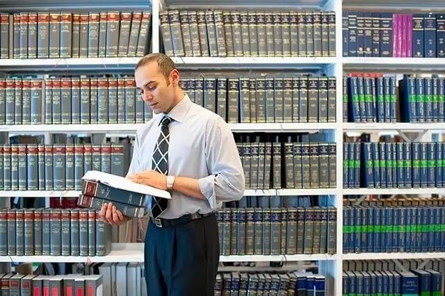 A man dressed in shirt and a tie in front of bookshelves, holding an open book and focused on reading.