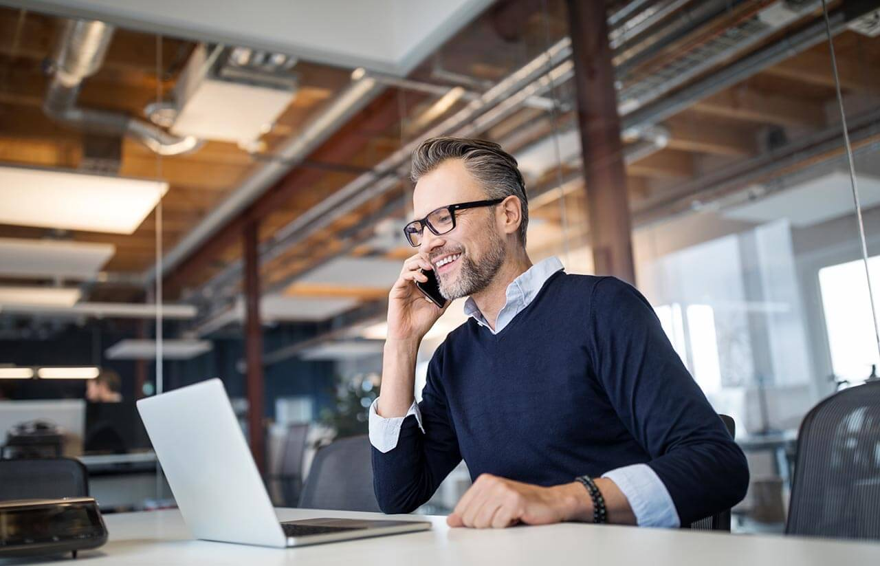 A man with glasses, wearing a navy sweater and white shirt, is sitting at a desk in an office. He is smiling while talking on a smartphone and using a laptop. The office has exposed pipes and beams in the ceiling.