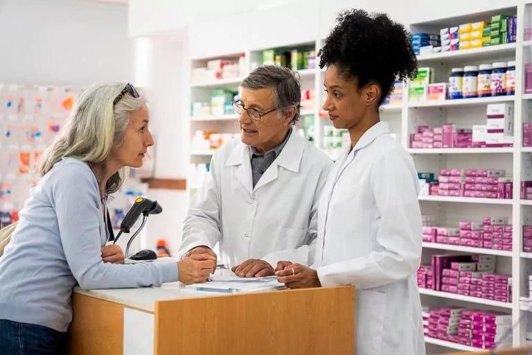 Woman consulting two pharmacists at a counter in a pharmacy, with health products visible behind glass cabinets.