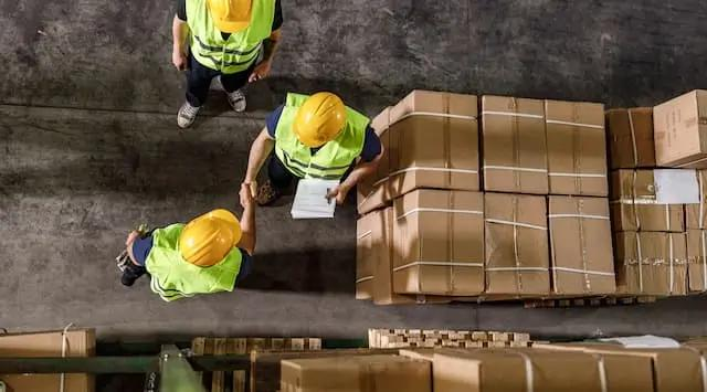 Three construction workers wearing yellow vests in a warehouse, discussing stock delivery.