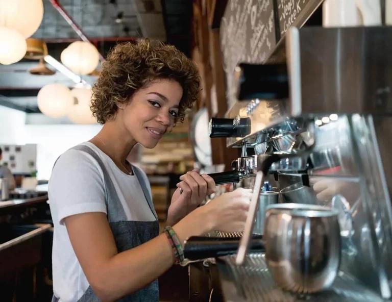 A woman prepares coffee behind the counter in a coffee shop.