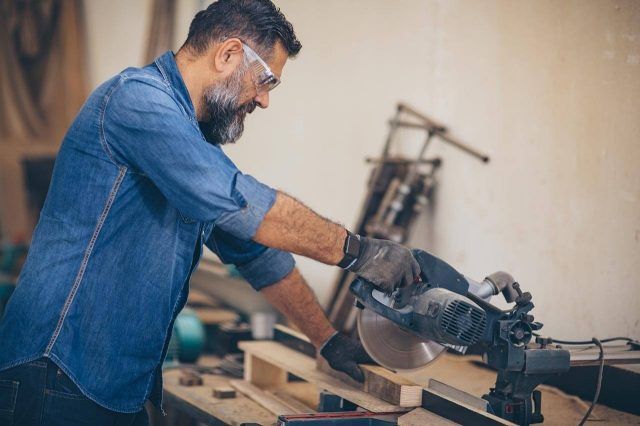 Wood worker using a miter saw to cut wood in a workshop, surrounded by tools.