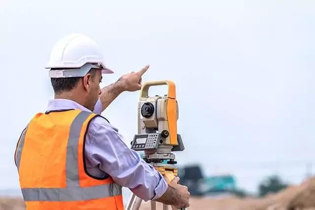Person in safety gear operating a surveyors level at a construction site under a clear sky.