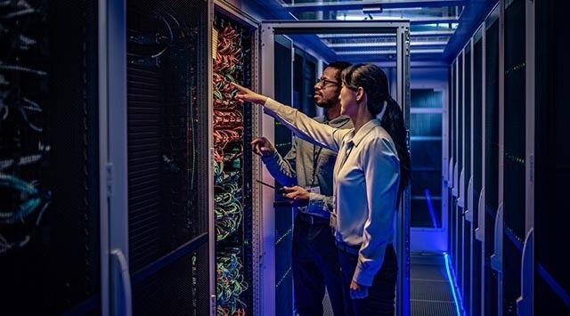 Two individuals stand in a dimly lit server room. They are examining a server rack filled with cables and equipment. One person points at the rack while both appear to be discussing or troubleshooting the setup.