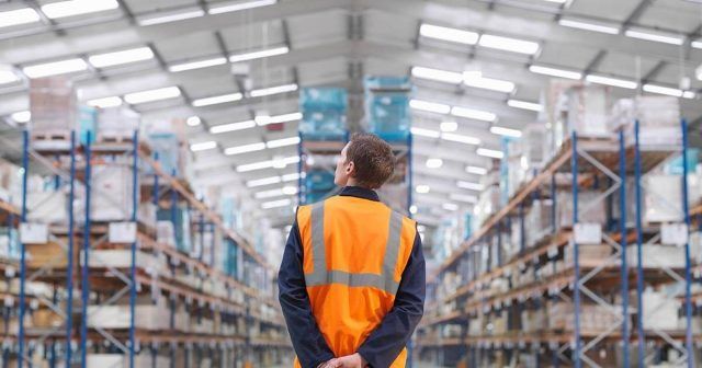 A person wearing an orange safety vest stands in a large warehouse, with shelves stacked high with boxes and pallets.