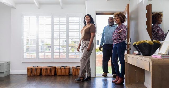 Three people are entering a home with a wooden floor; two women stand in the hallway while a man is at the door.