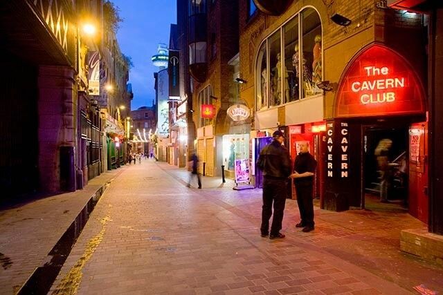 Two people near the entrance of The Cavern Club at dusk. The street is lit by neon and street lights.