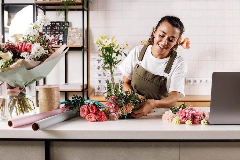 Florist arranging a bouquet on a counter, with gift wrapping materials and decorations in the background.