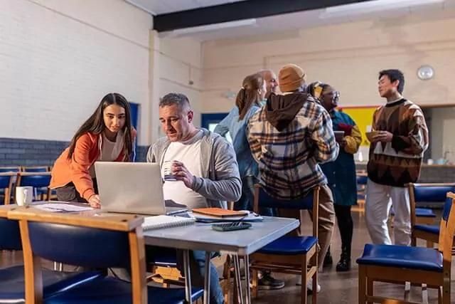 Man is working on a laptop at a table in a sports community centre while a woman is looking at the screen.