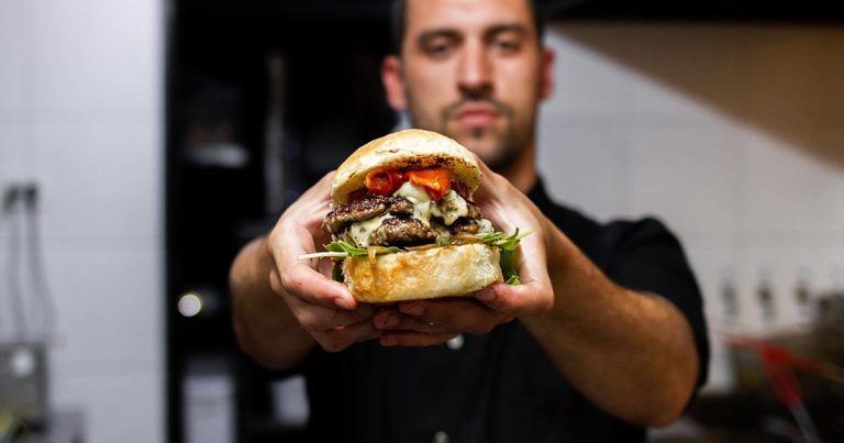Man holding a large sandwich in a kitchen with grilled meat, cheese, and vegetables between a bun, background blurred.