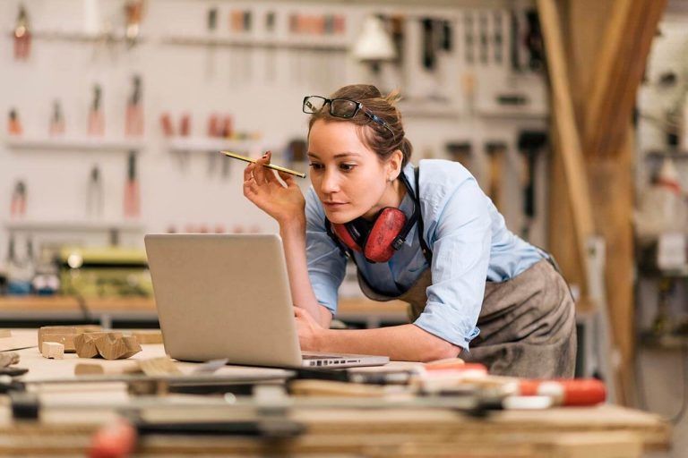 Woman working on a laptop in a workshop, holding a pencil with equipment and tools around her.