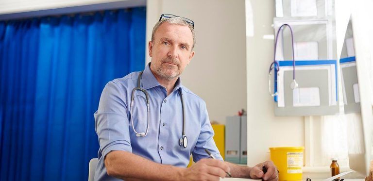 Doctor with stethoscope sitting at a desk in a medical office, glasses resting on his head.