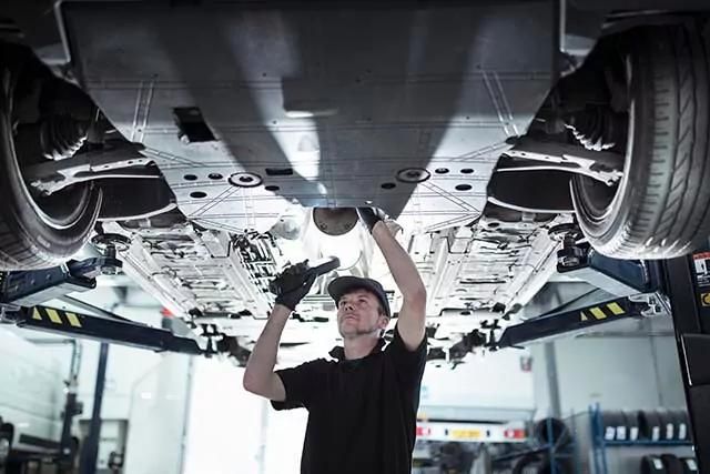 A mechanic is inspecting the underside of a vehicle raised on a hydraulic lift in a garage. He is wearing a cap, gloves, and a dark work shirt. The workshop environment is well-lit, with various tools and equipment in the background.