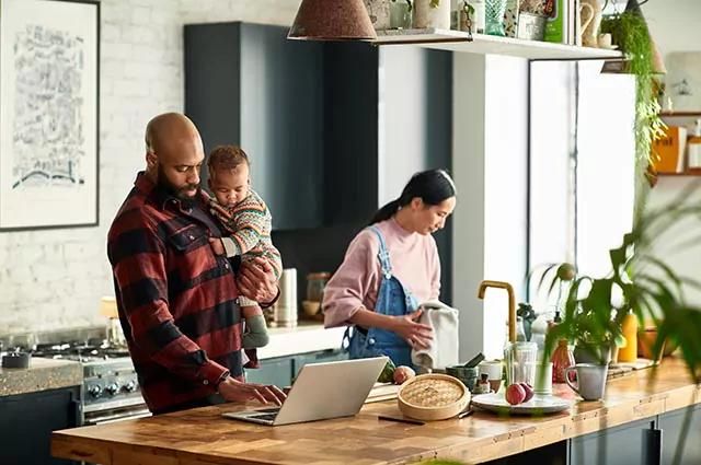 Man holding a child while using a laptop on a kitchen island, with a woman wiping her hands with a towel next to them.