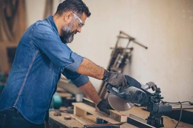 Wood worker using a miter saw to cut wood in a workshop, surrounded by tools.