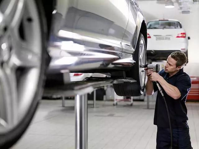 A mechanic works on a car elevated on a hydraulic lift, using a power tool to tighten or loosen bolts on a wheel. Another car is visible in the background, parked on an adjacent lift in a well-lit automotive repair shop.