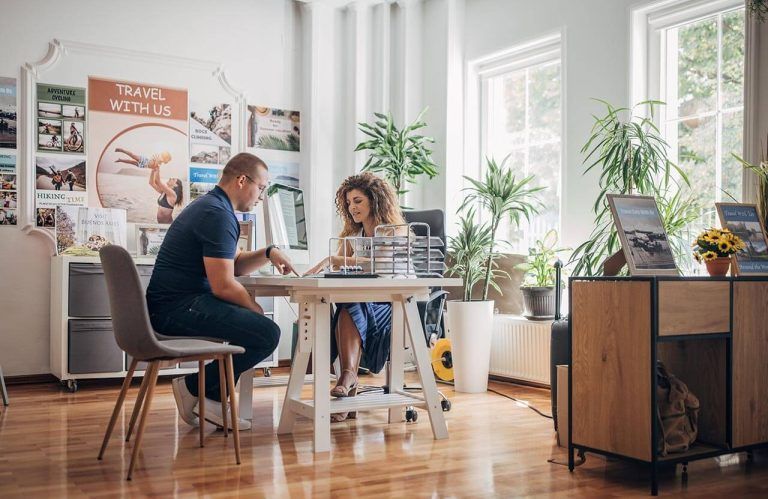 Man and woman at a travel agency desk, engaged in conversation, surrounded by travel posters.