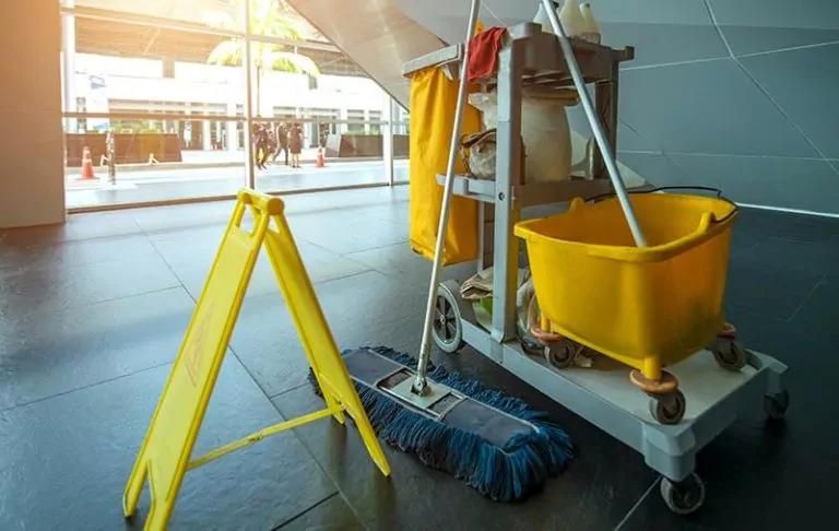 Cleaning trolley with mop and broom on a tiled floor with a caution sign nearby.