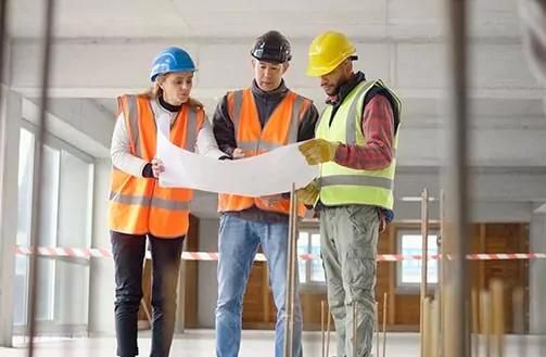 Three workers in safety vests and helmets examining a blueprint in an unfinished building.