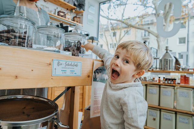Child with an astonished expression points at chocolate jars in a sweet shop.