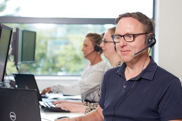 Smiling man with glasses wearing a headset and black zip-up jacket, looking at a laptop screen.