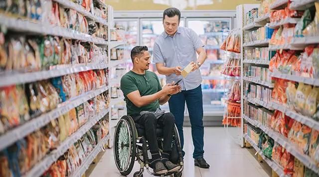Man in a wheelchair in a grocery aisle, assisted by another man holding a box.