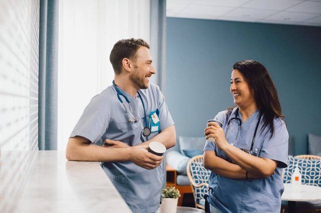Two healthcare professionals in scrubs smiling at each other with coffee cups in a break area with comfortable seating.