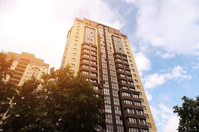 A tall residential apartment building, resembling a modern block of flats, stands against a partly cloudy sky with sunlight shining from the left. The building boasts numerous windows and geometric design elements, while trees with lush green leaves are seen at the bottom.