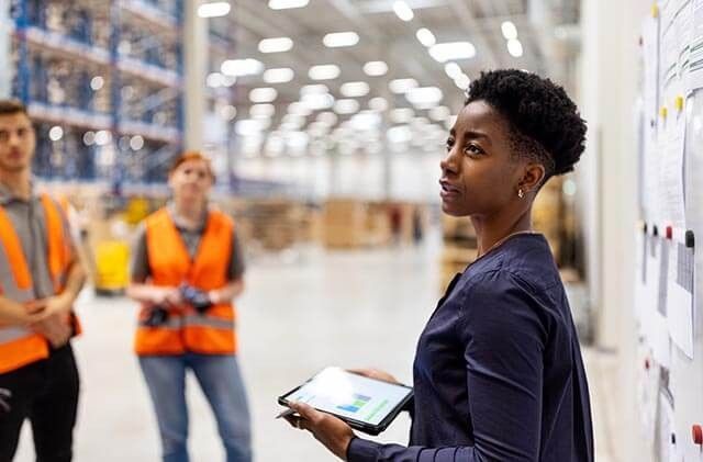 Person with tablet reviewing manufacturing processes in a warehouse, with two workers in the background.