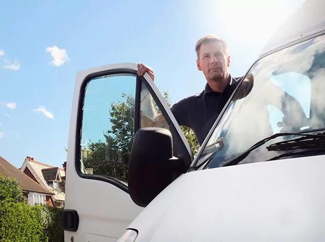 Person standing by an open van door under a sunny sky, with houses and greenery in the background.