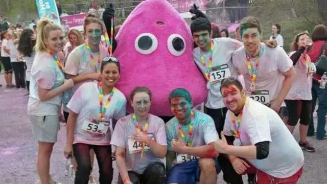 A group of people in colorful powder-stained outfits pose with a person in a large pink character costume. They are outdoors at a fun run event, wearing race numbers and rainbow lanyards. The ground is dusted with colored powder.